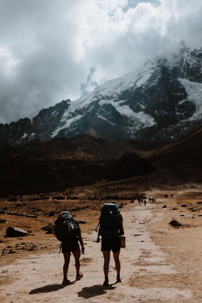 Two hikers with backpacks walk on a rugged path towards a snow-covered mountain under a cloudy sky.